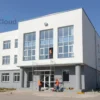 A modern hospital building in Chelyabinsk region, Russia, with construction workers repairing the exterior. The hospital has a clean, well-maintained facade with large windows. The workers are using scaffolding and various tools. The scene is set during the day with clear blue skies and sunlight illuminating the building.