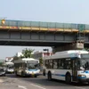 A bustling urban scene showing a large bridge under construction with scaffolding and safety barriers, while several city buses travel on a nearby alternate route marked by a simple arrow sign, all under a clear sky; the focus is on the movement of the buses and the partially repaired bridge without any visible text or logos