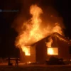 A burning house in a small village in Sverdlovsk Oblast, flames engulfing the wooden structure, thick smoke rising into the night sky, dark silhouettes of nearby trees, a sense of urgency and destruction, realistic depiction of fire and smoke