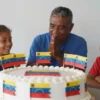 A large, colorful cake with Venezuelan flags and children's faces decorated on it. The children are smiling and looking at the cake with joy. The background shows a simple, clean environment suggesting a shelter. Focus on the children's expressions of happiness and the celebratory nature of the cake.