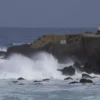 A dramatic coastal scene in Kumamoto, Japan showing powerful ocean waves crashing against rugged cliffs with visible seismic activity like tremors or minor rockfalls, distant tsunami warning signs or evacuation boats on the horizon, dark stormy sky with lightning, realistic ocean details, no text or numbers on image