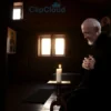 A serene elderly Orthodox monk in traditional robes kneeling in quiet prayer at dawn inside a modest wooden chapel, soft golden light filtering through small windows, icons on the walls, a single lit candle on a wooden stand, hands folded in reverence, peaceful expression, no text or symbols visible