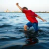 A 10-year-old boy in a red shirt and blue shorts struggling in the Volga River, surrounded by calm water, with a distant cityscape of Volgograd on the horizon, dramatic lighting, emotional atmosphere
