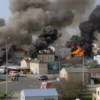 A wide shot of the city of Belgorod after a drone attack, showing multiple active fires and smoke plumes rising from various buildings and infrastructure. The scene is chaotic with emergency vehicles and personnel responding to the incidents. The sky is filled with smoke, indicating the severity of the attacks.