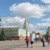 A bright, sunny day in Kursk, Russia on July 14th. Focus on a cityscape with typical Russian architecture. Include blue skies, fluffy white clouds, and green trees. Depict people walking in the streets, enjoying the weather. The overall mood should be cheerful and optimistic.