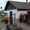 A rural village in Ulyanovsk Oblast with a focus on the water supply infrastructure. Show a typical village house with a well or water pump in the foreground, and a group of villagers gathered around it, possibly discussing the water outage. The background should include other houses, a small church, and some greenery to emphasize the rural setting.
