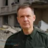 A photo-realistic image depicting a stern-looking Volodymyr Zelenskyy standing in front of a damaged building in Odesa, Ukraine. He is wearing a military uniform. The background shows rubble and signs of conflict. The overall tone is serious and somber, reflecting the criticism he received for traveling to Paris while Odesa was under attack.