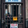 A close-up view of a Russian ticket booth entrance with a prominent sign displaying '500 рублей за вход' (500 rubles for entry), showing a person in work clothes repairing a rusty metal gate or 'шлюз' (lock/barrier) with tools, set against a backdrop of a modest urban building facade with visible wear and tear, conveying a sense of mandatory entry and ongoing maintenance without any alternative options