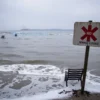 A bleak, polluted beach scene in Nagaria where the sea is closed due to sewage contamination. Dark, murky water reaches the shoreline, with foam and debris floating. A rusted, warning sign with a red cross (no text visible) stands near the water's edge. Overcast sky, distant industrial silhouettes, and a few abandoned beach chairs half-submerged. The atmosphere is somber and emphasizes environmental damage.