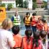 A group of children in Smolensk gathered near a calm riverbank or pool, attentively listening to a safety instructor demonstrating water safety practices. The scene includes bright life jackets, safety signs, and a rescue float, with the instructor showing proper techniques. The children are diverse, wearing casual summer clothing, and the background shows typical Russian architecture or natural scenery, emphasizing a safe and educational environment.