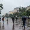 A quiet weekend morning in Chelyabinsk region with light rain falling over a small cityscape, wet streets reflecting gray skies, people with umbrellas walking slowly, trees glistening with raindrops, no bright colors, calm and peaceful atmosphere