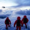 A dramatic mountain rescue scene on Mount Elbrus at dawn, showing two rescue teams in bright orange gear searching through deep snow and icy terrain for four missing climbers, with a helicopter hovering overhead and stormy clouds in the background, conveying urgency and harsh conditions