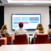 A group of people attending a meeting in a conference room. The room is well-lit with modern furniture. A large screen displays data or a presentation. Focus on the people engaged in discussion.