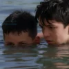 Two boys swimming in a river, one boy is submerged underwater with only his head visible above the surface, the other boy is floating on his back looking worried, calm river water, natural lighting, realistic style