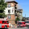 A residential building, two stories tall, with visible damage from an explosion. Debris and smoke are present around the building. Emergency vehicles (fire trucks, ambulances) are on the scene. The scene depicts a daytime setting. Focus on the aftermath and the building's structural damage.