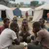 A somber scene depicting a refugee camp in the Democratic Republic of Congo, with people huddled together, displaying signs of distress and illness. Focus on conveying the humanitarian crisis and the devastating impact of the Ebola outbreak.