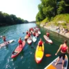 A wide shot of a group of people paddling stand-up paddleboards (SUPs) on the Dnipro River in Smolensk, Russia. The scene is vibrant with summer activity, featuring clear blue water, green banks lined with trees, and sunny skies. The people are wearing brightly colored clothing and life vests. Several SUPs are close together, creating a sense of community and shared activity.