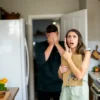 A food blogger in her kitchen, looking shocked and surprised, with a delivery person standing in the doorway, appearing embarrassed and covering himself with his hands. The kitchen is well-lit and filled with cooking utensils and food ingredients.