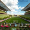 A wide shot of the Spartakovec stadium undergoing landscaping. The stadium is modern with green grass, trees, and flowerbeds surrounding the field. People are working on planting trees and maintaining the greenery. The sky is blue with some clouds.