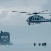 A dramatic scene showing a helicopter from the Russian Emergency Situations Ministry (MCHS) hovering above a remote Arctic coastline, with tourists disappearing into the distance on the ground, conveying a sense of urgency and isolation