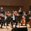 A vibrant, medium shot of the Smolensk UFSIN orchestra performing on a stage. The orchestra consists of various musicians playing instruments like violins, cellos, trumpets, and drums. The stage is well-lit with spotlights, and the musicians are dressed in formal attire. In the background, a blurred audience can be seen. The overall atmosphere is celebratory and professional.