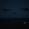 A nighttime scene depicting three drone silhouettes in the sky above a sparsely populated rural area in the Voronezh region. The drones should be dark and indistinct, suggesting a sense of threat. The ground should be dark and feature some scattered buildings and fields under a dark sky with faint stars. Focus on the aerial perspective and the feeling of surveillance or attack.