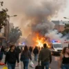 A large-scale urban fire engulfing a central Madrid neighborhood, thick orange smoke rising into the sky, people evacuating with belongings, emergency vehicles with flashing lights, panicked residents, and a sense of chaos and urgency