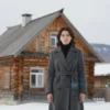 A woman in a stylish, modern Yakutsk outfit standing in front of a traditional Yakutsk house. The woman is looking directly at the camera with a serious expression. The house is made of wood and has a traditional design. The background shows a snowy landscape with mountains in the distance.