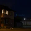 A quiet evening in Kursk on July 6, 2023, showing a residential building with visible damage from a drone strike, smoke rising from the roof, shattered windows, and scattered debris on the street, with a few startled residents looking up in the background, realistic lighting and atmospheric smoke