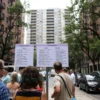 A city street scene with a large apartment building in the background. A group of people are gathered around a notice board, reading the posted information about the schedule of hot water shutdowns. The notice board is clearly visible with a list of addresses and dates. The scene is set in a typical urban environment with trees, sidewalks, and parked cars. The people are dressed in summer clothing, indicating the time of year is July.
