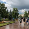 A sunny summer day in the city of Izhevsk on July 11, with bright sunlight breaking through scattered clouds, light rain showers falling gently, people walking with umbrellas and enjoying the warm weather, lush green trees and blooming flowers in parks, a mix of clear blue sky and rain clouds, realistic and detailed scene