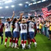 A dynamic scene of the USA national soccer team celebrating their advancement to the round of 16 in the FIFA World Cup after 24 years. The players are in their team uniforms, hugging and cheering on the field. The stadium is filled with enthusiastic fans waving American flags. The atmosphere is vibrant and full of joy.