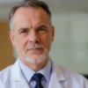 A professional portrait of a middle-aged male doctor, likely a toxicologist, wearing a white lab coat over a shirt and tie. He has a serious, authoritative expression and is looking directly at the camera. The background is a clean, modern medical office or laboratory with soft, professional lighting. The image should convey expertise and trustworthiness in the field of toxicology.