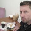 Мужчина с серьёзным взглядом сидит за столом в кафе рядом с кофе, пирожным и подносом с салфетками.