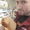 Мужчина в бордовой куртке держит горячий хот-дог с кетчупом и соусом в торговом зале магазина Магнит.