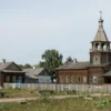 A peaceful rural landscape in Yaroslavl region showing a historic wooden church or village square with subtle signs of vandalism such as graffiti on a fence or broken windows, but overall calm atmosphere, no people, no text, no letters or digits