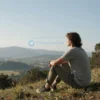 A serene and contemplative scene with a person sitting on a hillside, gazing at a vast landscape under a clear sky. The person is in a relaxed posture, surrounded by nature, with mountains in the distance and a few trees nearby. The atmosphere is peaceful and reflective.