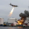 A military aircraft releasing bombs over a Ukrainian port and fuel storage facility, with explosions and smoke rising from damaged infrastructure, surrounded by industrial buildings and cargo containers, under a cloudy sky