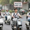 A group of young people riding electric scooters on a city street, one of them holding a traffic sign that reads 'Know the traffic rules'