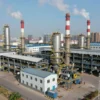 A modern industrial factory complex with smokestacks emitting smoke, surrounded by a cityscape with buildings and roads. Include workers in uniforms operating machinery inside the factory, and a clear sky above.