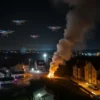 A nighttime aerial view of a residential area in Tula Oblast under attack, with multiple drones flying in formation, illuminating the sky with lights, and causing visible damage to houses and infrastructure below. Smoke rises from burning buildings, emergency vehicles are present, and the scene is depicted with realistic detail and dramatic lighting.