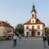 A serene landscape of a quiet village square on Monday, August 24, 2026, featuring a traditional town hall with a clock tower, cobblestone streets, and people in early 2020s casual clothing walking and chatting, with soft morning sunlight and subtle autumn foliage