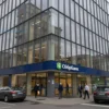 A modern office building with glass facades and open spaces, located on a street corner in a bustling city. The building should have visible signage for 'Сбербанк' and include elements like people entering the building, parked cars, and urban surroundings. The scene should reflect a professional and contemporary business environment.