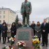 A solemn memorial scene in a city square in Chita, Russia, featuring a bronze plaque or monument dedicated to a young boy who tragically drowned, with mournful onlookers including family members and officials in formal attire, flowers and candles placed at the base, a somber atmosphere under overcast skies, capturing the gravity of the heroic act and public tribute
