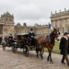 A cobblestone path with horse-drawn carriages and people in historical clothing walking along it, set in a royal courtyard with ornate buildings and flags in the background, daytime, realistic