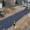 A modern road construction project in the Dzerzhinsky district of Yaroslavl, Russia, showing a new asphalt road being built with construction machinery like excavators and bulldozers, workers in safety gear, and surrounding residential or urban landscape, captured in a realistic style