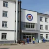 A modern cadet school building in Primorsky District with visible water pipes being replaced, construction workers in uniform, school emblem on facade, bright daylight, urban street setting, no text or numbers on image