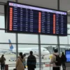 A modern airport terminal building with a large digital departure board showing flight information, airplanes on the tarmac, and travelers with luggage in the foreground, conveying a sense of halted operations