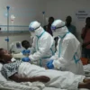 A somber scene depicting a crowded hospital ward in the Democratic Republic of Congo, with medical staff in protective gear attending to patients lying on beds, some with IV drips, while others are being monitored. The atmosphere is tense, with dim lighting and a sense of urgency. Include a few individuals in the background showing signs of distress, and a sign indicating 'Ebola Treatment Center' in the background.