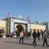 A festive pedestrian center in Ryazan opened during the Day of the City celebration. The scene includes a decorated entrance with banners, flags, and floral arrangements. People are walking, smiling, and enjoying the event. There are colorful lights, balloons, and a welcoming atmosphere. The architecture is modern with traditional elements, and the sky is clear and bright.